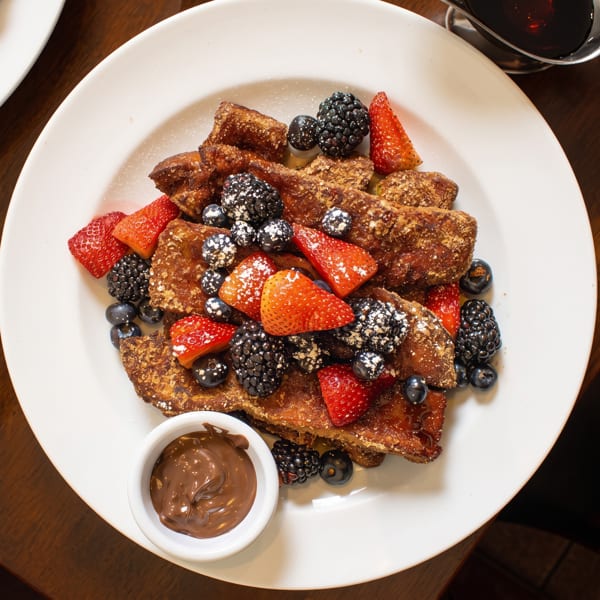 French toast topped with strawberries, blueberries, blackberries, powdered sugar, and chocolate sauce on a white plate.