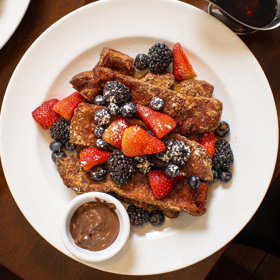French toast topped with strawberries, blueberries, blackberries, powdered sugar, and chocolate sauce on a white plate.