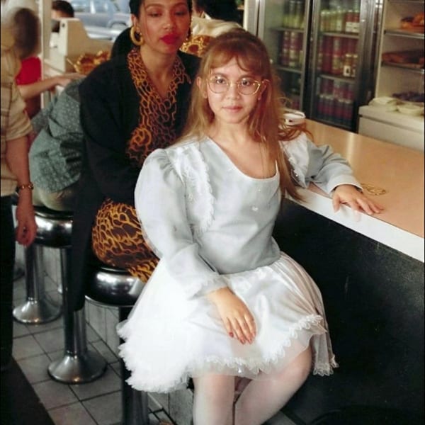 Two women pose at a restaurant counter, with stools, a refrigerated drink case, and other diners visible behind them.