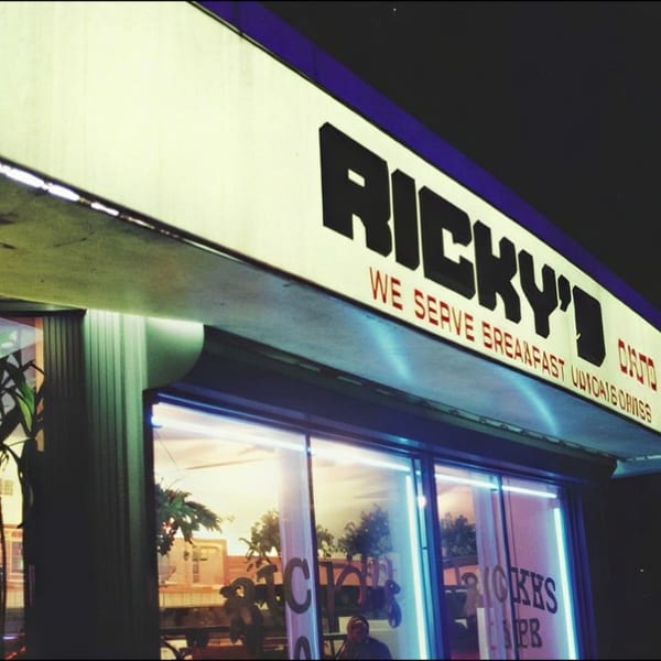 Nighttime exterior of Ricky’s diner, with illuminated signage advertising breakfast, lunch, dinner, and burgers.
