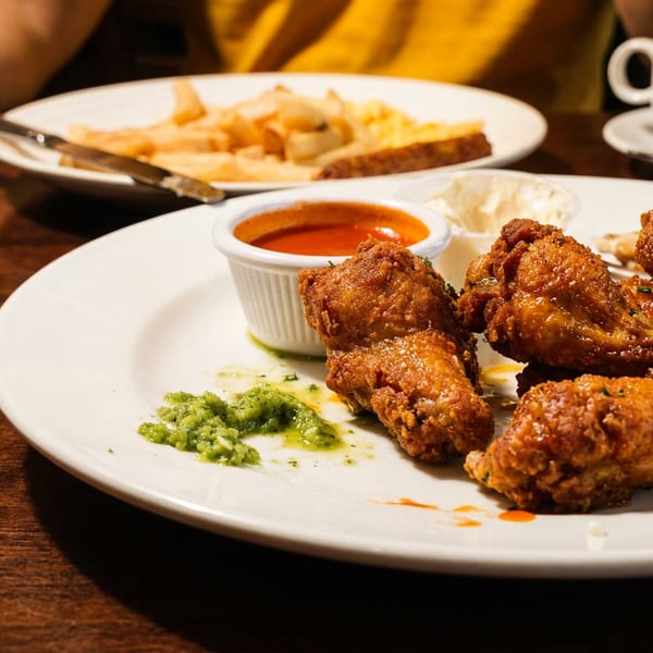 Crispy fried chicken pieces with dipping sauces on a white plate, accompanied by fries, coffee, and a diner in the background.