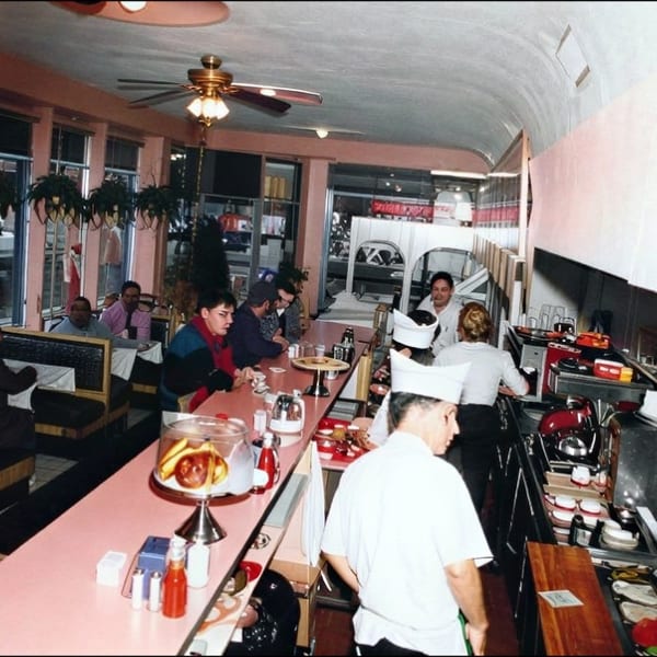 Busy retro diner with pink counters, seated patrons, hanging plants, and staff preparing food behind the counter.