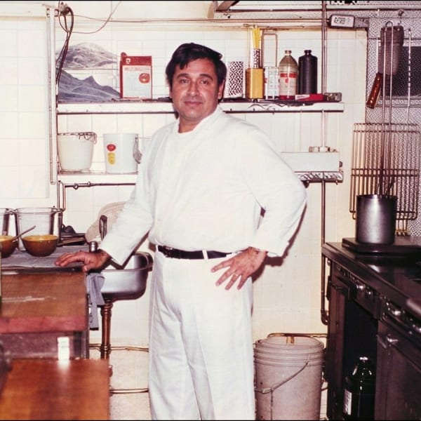 Chef in white uniform stands in a vintage restaurant kitchen surrounded by shelves, cookware, bottles, and a large stove.