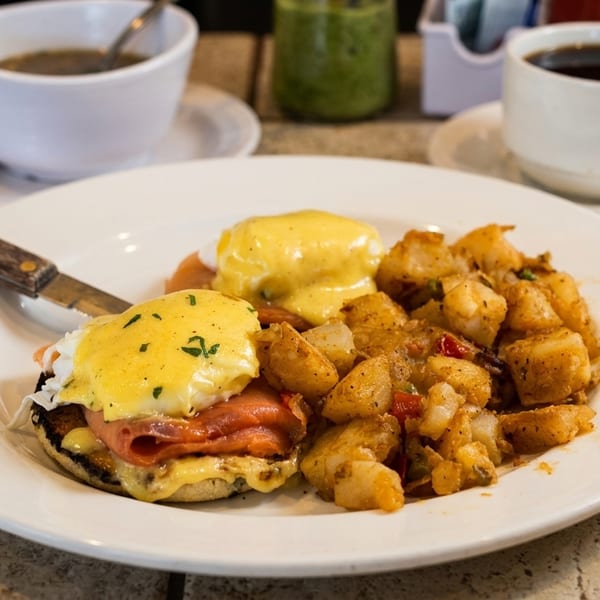 Eggs Benedict with ham and hollandaise, served with seasoned breakfast potatoes, alongside two cups of coffee.