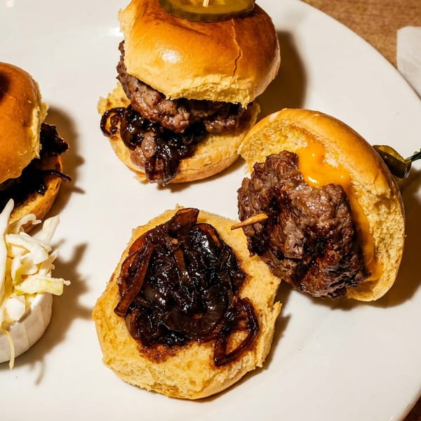 Three gourmet sliders with pickles, caramelized onions, melted cheese, and coleslaw arranged on a white plate.