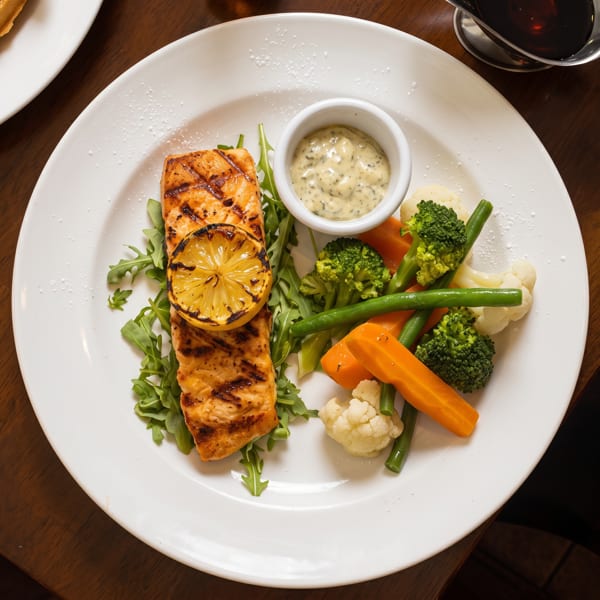 Grilled salmon topped with charred lemon, mixed vegetables, greens, and creamy sauce on a white plate.