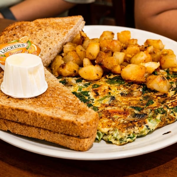 Plate of spinach omelet, roasted potatoes, toasted wheat bread, butter, and jam on a wooden restaurant table.