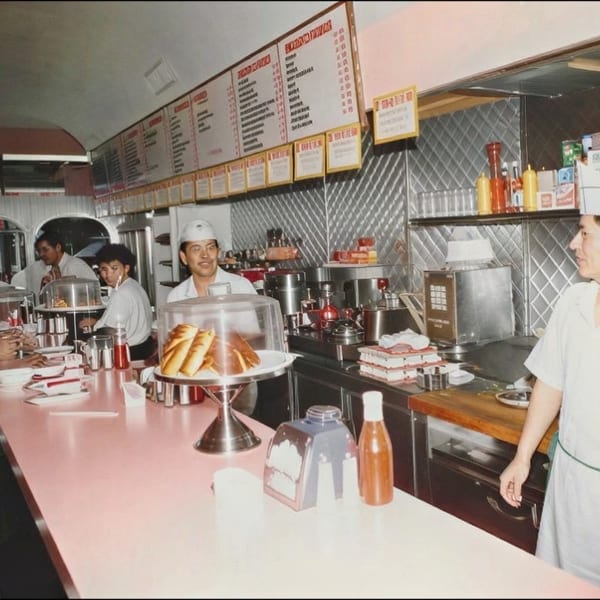 Vintage diner interior with customers at a counter, staff in white uniforms, menu boards, condiments, and pastries displayed.