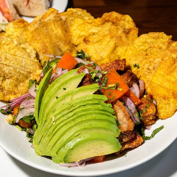 Golden fried plantains surround avocado, seasoned meat, tomatoes, onions, and herbs on a white plate.