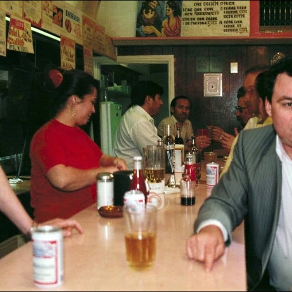 Busy traditional restaurant counter with diners, drinks, condiments, handwritten signs, and a server in a red shirt.