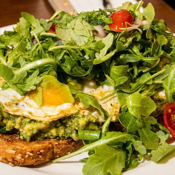 Avocado toast topped with a fried egg, arugula, cherry tomatoes, and greens on a white plate.