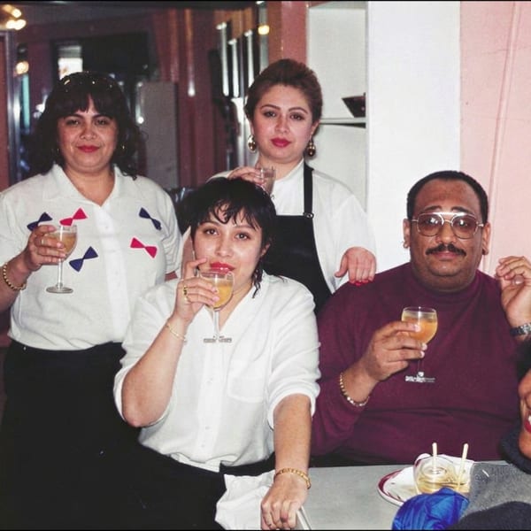 Five people smile and raise drinks together inside a warmly lit restaurant.