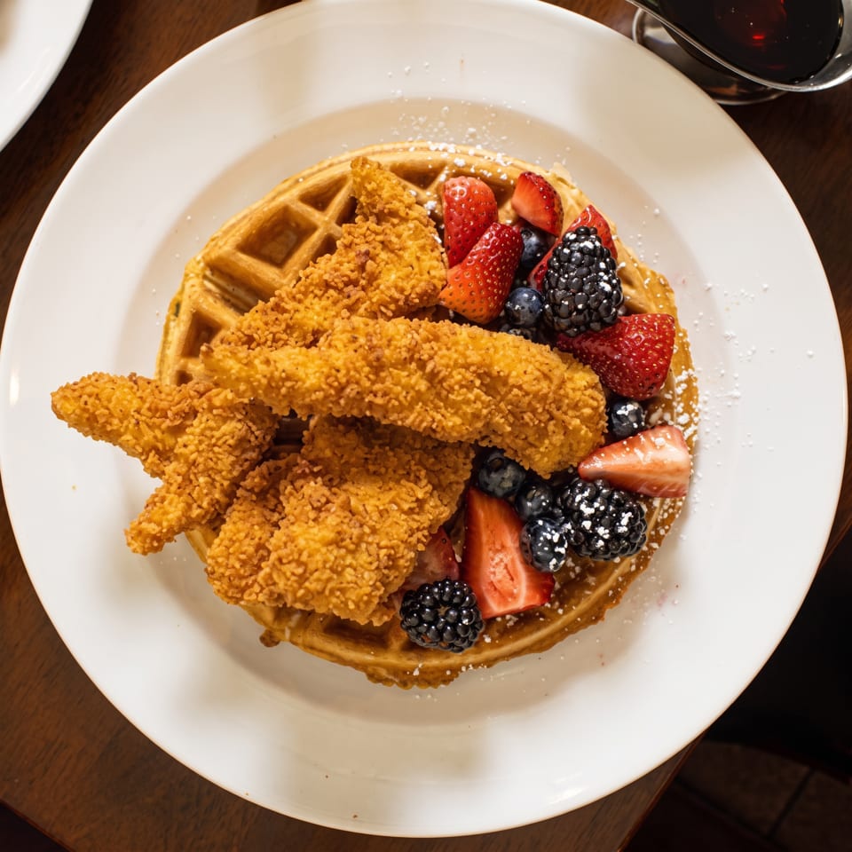 Golden waffle topped with crispy chicken tenders, strawberries, blueberries, blackberries, and powdered sugar.