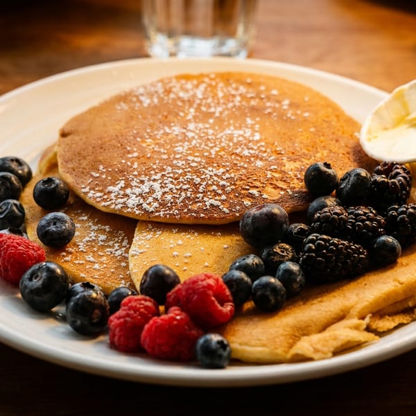Stack of powdered-sugar pancakes topped with blueberries, raspberries, blackberries, and a cup of cream on a white plate.