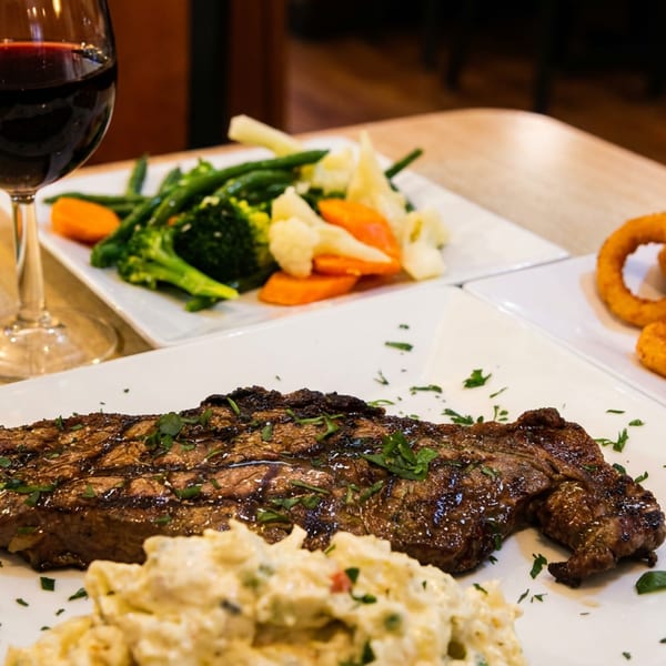 Grilled steak garnished with herbs, creamy side dish, vegetables, onion rings, and red wine on a restaurant table.