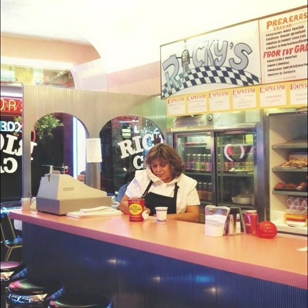 Diner counter with a server, chrome stools, refrigerated drinks, and RICKY’S signage advertising burgers and crab.